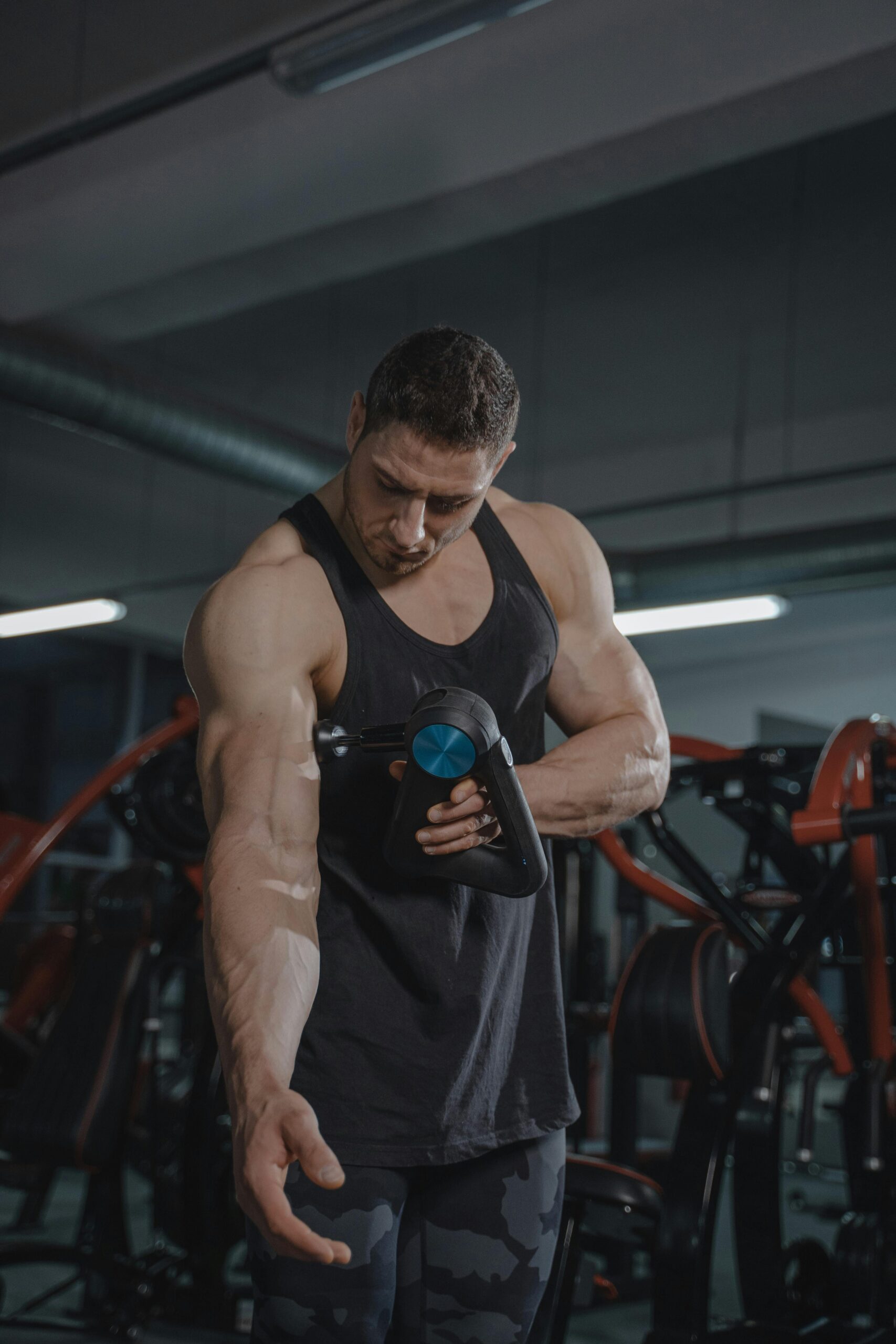Man in activewear massaging arm with a massage gun in a gym, highlighting muscle definition.